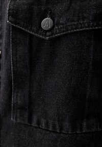 Szczegół czarnej kurtki dżinsowej z guzikiem z wytłoczonym logo, kontrastowym przeszyciem i teksturowanym materiałem. Zbliżenie na kieszeń na piersi.