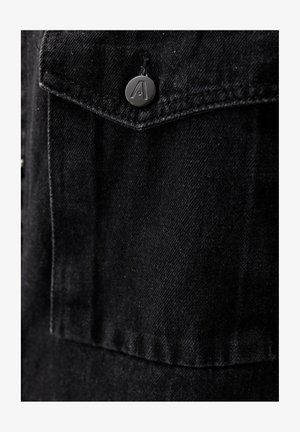 Szczegół czarnej kurtki dżinsowej z guzikiem z wytłoczonym logo, kontrastowym przeszyciem i teksturowanym materiałem. Zbliżenie na kieszeń na piersi.