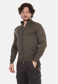 Maglione zip-up verde scuro con colletto alto, design aderente, texture a coste e cuciture piatte, indossato sopra pantaloni cargo scuri.