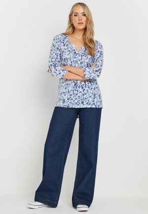 Bloemenprint top in lichtblauwe tinten met ronde hals en driekwartmouwen; gecombineerd met wijde donkere spijkerbroek en witte sneakers.