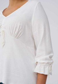 Witte blouse met een V-hals en geplooid detail bij de buste. Heeft driekwartmouwen met gerimpelde manchetten en trekkoordaccenten.