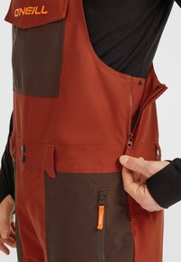 Pantalones de peto rojos y marrones hechos de tejido resistente al agua, con múltiples bolsillos, un bolsillo en el pecho y un bolsillo lateral con cremallera que tiene una lengüeta naranja.
