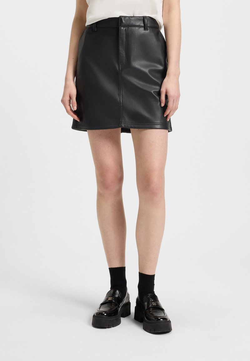 Jupe mini en simili cuir noir avec une couture frontale et une taille, tombant au-dessus du genou ; associée à des chaussures à plateforme noires et chunky.
