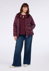 Kvinna stående med händerna i fickorna, iförd en lila pufferjacka, matchande tröja med utskärningar, vida blå jeans och vita sneakers.