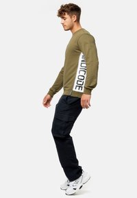 Sweatshirt vert olive avec bande latérale blanche comportant un texte noir. Associé à un pantalon cargo noir et des baskets blanches. Look décontracté et sportif.