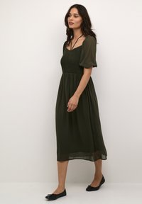 Rochie midi verde măsliniu cu un bust elastic, mâneci scurte pufoase și o fustă fluidă. Asortată cu pantofi plata negri.