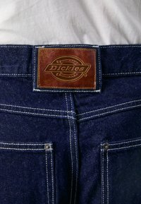 Džinsai su rudu odiniu ženkleliu, ant kurio yra "Dickies" logotipas, kontrastinėmis baltomis siūlėmis ir sidabro spalvos metaliniais smeigėmis ant galinių kišenių.