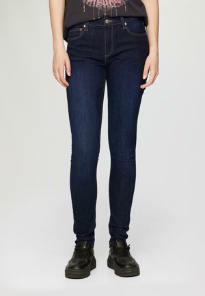 Skinny denim jeans in dark blue, featuring a classic mid-rise waist, five-pocket design, and subtle stitching accents. Rubber-soled black shoes.