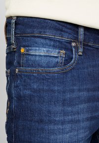 Vaqueros de mezclilla azul oscuro con un corte ajustado, con costuras amarillas, un pequeño bolsillo frontal y un detalle de botón metálico en la cintura.