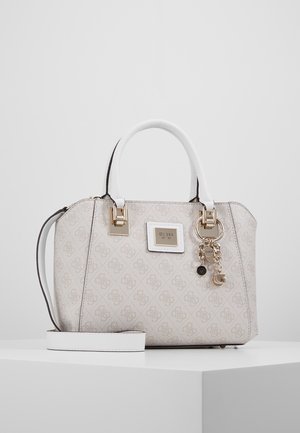White Guess handbag with monogram pattern, double handles, gold hardware, logo plate, and gold charm details, resting on a white surface.