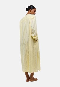 Robe jaune, longue, en tissu léger et transparent avec des rayures verticales, présentant un dos froncé, des manches longues et des liens à la taille. Tissu texturé léger.