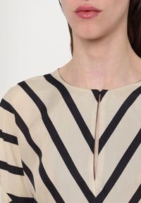 Gros plan d'une personne portant un haut beige avec de larges rayures noires diagonales et horizontales et une petite fente à l'encolure devant.