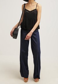 Woman wearing black sleeveless top, navy wide-leg pants, tan platform sandals, and carrying a black round crossbody bag.