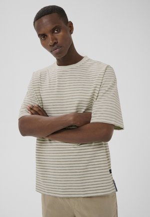 Mand med krydsede arme iført beige kortærmet skjorte med striber og lysebrune bukser foran ensfarvet baggrund.