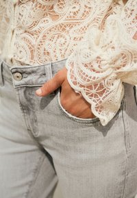 Top bianco in pizzo a maniche lunghe con motivi intricati abbinato a jeans grigio chiaro. La mano è poggiata nella tasca anteriore dei jeans.