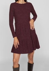 Burgundské páskované šaty s dlouhými rukávy, kulatým výstřihem a rozšířenou sukní; tkanina s texturou a sklady v oblasti pasu.