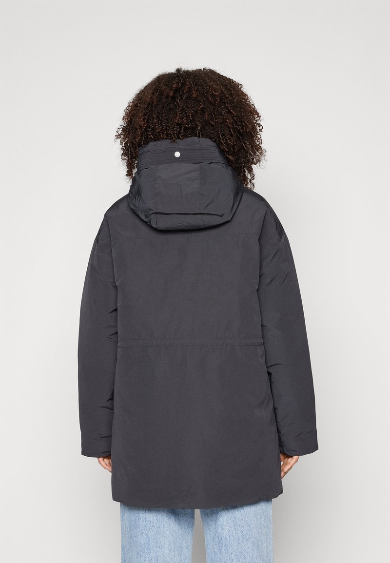 Veste isolante noire avec capuche, présentant une texture lisse et un design mi-long, associée à un jean bleu clair.