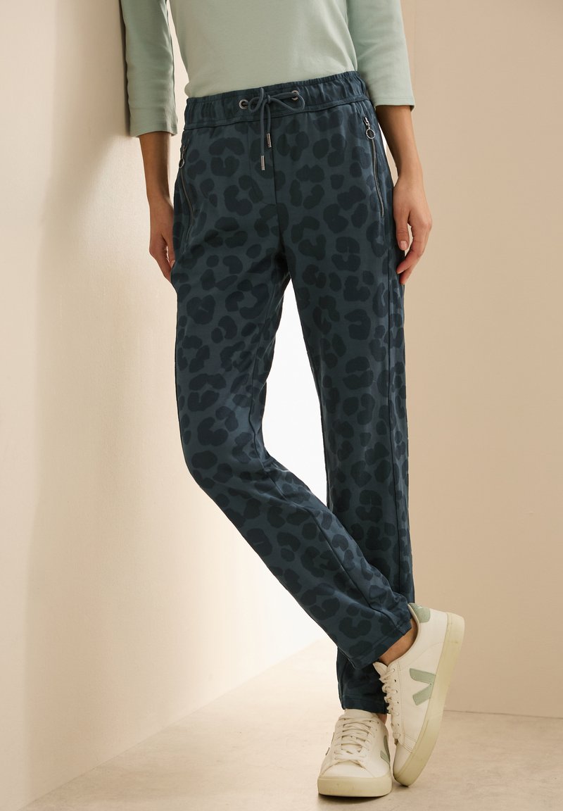 Leopardmönstrade joggers i mörk teal, tillverkade av mjukt tyg; har dragsko i midjan och dragkedjefickor på sidan. Matchat med vita sneakers.