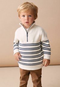 Suéter blanco y azul marino a rayas con media cremallera y textura de canalé, combinado con pantalones marrones. Niño de pie con una expresión neutral.