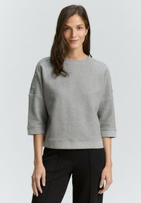 Grauer Sweatshirt mit Textur, Rundhalsausschnitt und Dreiviertelärmel, mit Knopfdetails an den Schultern. Getragen mit schwarzen Hosen.