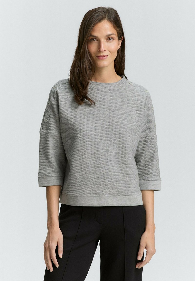 Grauer Sweatshirt mit Textur, Rundhalsausschnitt und Dreiviertelärmel, mit Knopfdetails an den Schultern. Getragen mit schwarzen Hosen.