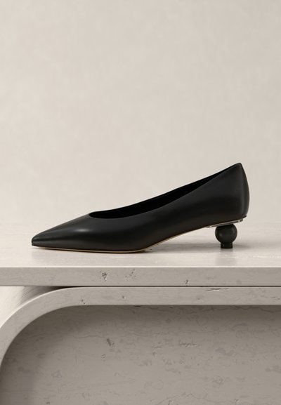 Zapato de mujer de cuero negro con tacón bajo, punta puntiaguda y talón redondeado y esculpido, exhibido sobre una superficie de piedra beige clara.