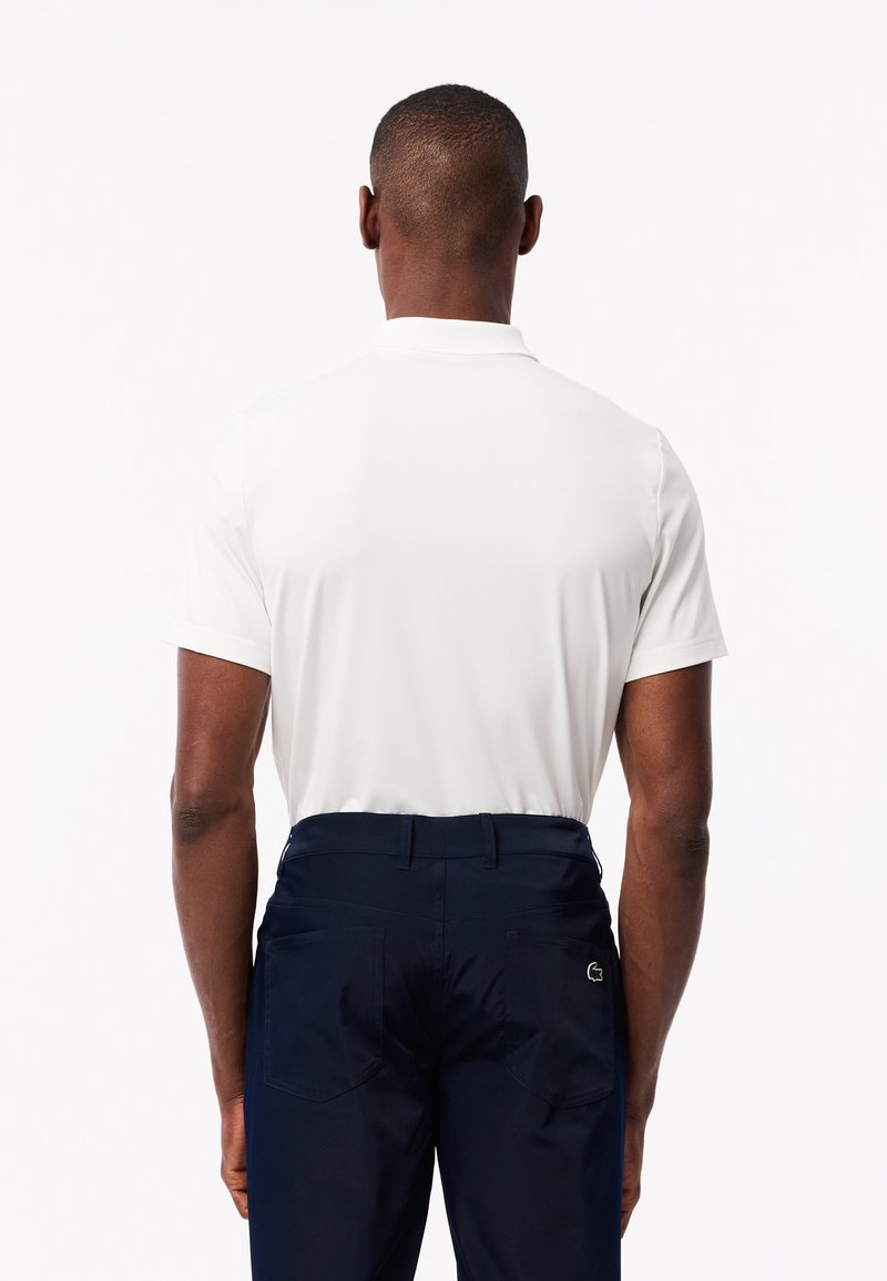 Polo shirt blanc à manches courtes avec un col, associé à un pantalon bleu marine avec des poches arrière et un petit logo. Texture lisse.