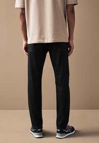 Schwarze Jeans mit geradem Bein, kombiniert mit einem beigefarbenen T-Shirt mit kurzen Ärmeln. Der Stoff wirkt glatt und mit wenigen Details.