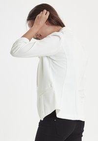 Blazer bianco con vestibilità su misura, tessuto morbido, schiena allungata e due tasche frontali. Presenta un classico colletto a punta e maniche arricciate.