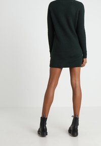Vestido de punto verde oscuro con mangas largas y cuello redondo, con textura acanalada y una silueta ceñida. Botas negras hasta el tobillo.