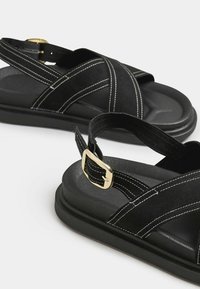 Mustat sandaalit, joissa on ristikkäiset hihnat, valkoiset ompeleet, säädettävä nilkkahihna ja kultainen solki teksturoidulla mustalla pohjalla.