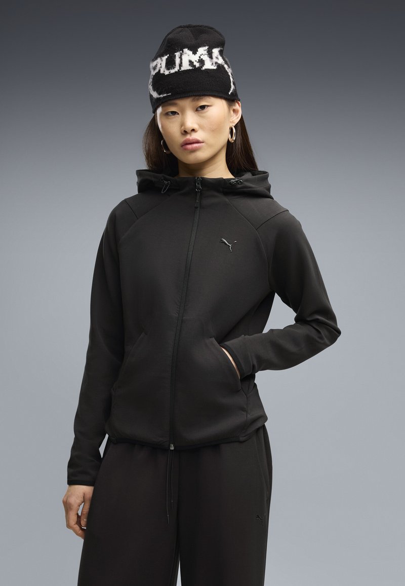 Femme portant un sweat à capuche zippé noir Puma, un pantalon noir assorti, un bonnet noir Puma et des boucles d'oreilles créoles sur fond gris.