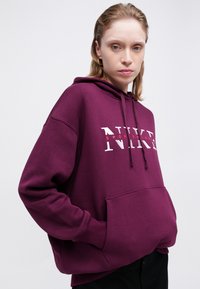 Vijolična kapuca iz mehkega blaga z sprednjim žepom. Ima velik bel logotip "NIKE" in manjši rdeč napis "sportswear" spodaj.