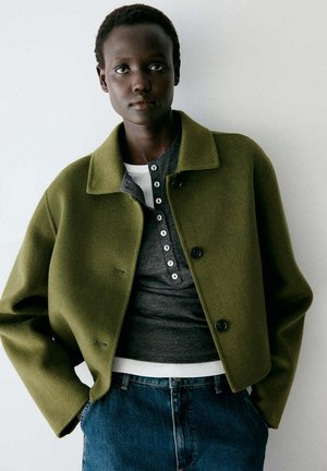 Persona con pelo corto que lleva una chaqueta verde oliva abotonada sobre una camisa gris oscuro y blanca y pantalones vaqueros azules, de pie frente a un fondo liso.