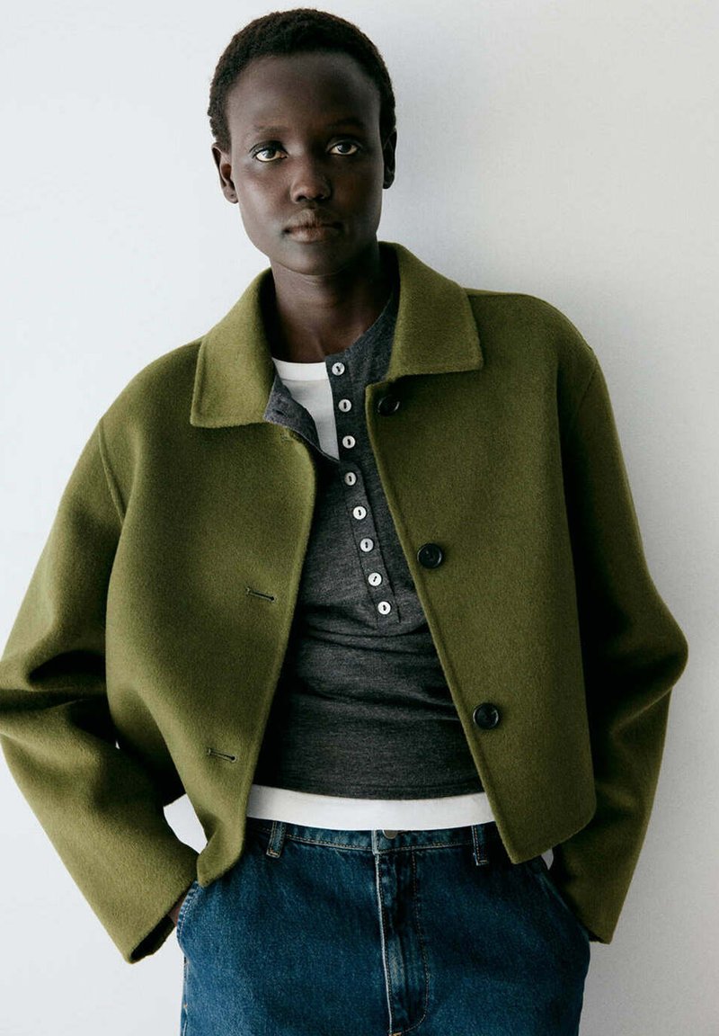 Personne aux cheveux courts portant une veste boutonnée vert olive sur une chemise gris foncé et blanche ainsi qu'un jean bleu, debout devant un fond uni.