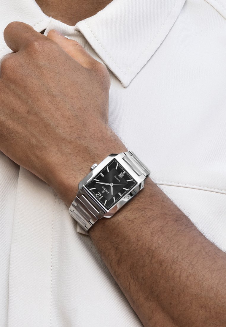 Srebrn metalni ročni ura s pravokotnim črnim številčnikom, srebrnimi kazalci in poliranim zapestjem. Odpeta na roki ob belem srajcu.