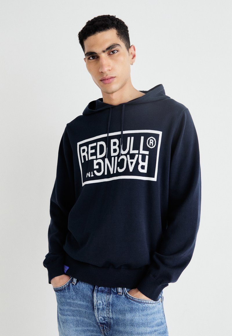 Ungdommelig mann med kort, krøllete hår iført en marineblå hettegenser med teksten "RED BULL RACING" speilvendt, blå jeans, står mot en hvit bakgrunn.