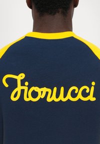 Bakgrunnsvisning av en person som har på seg en marineblå og gul skjorte med ordet "Fiorucci" skrevet med gult på den øvre delen av ryggen.