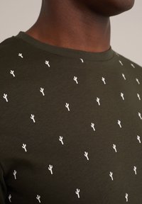 Camiseta verde oliva con estampado de cactus blancos dispersos. Cuello redondo, textura de tejido simple y mangas cortas.