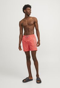 Bañador de coral con cinturilla elástica y cordón gris. Presenta un logo en texto blanco en el lado izquierdo. Se lleva con sandalias de piscina negras.