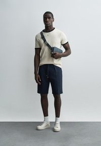 Ung mand iført beige T-shirt med sorte kanter, marineblå shorts, hvide sneakers, hvide sokker og en sort skuldertaske, står op ad en ensfarvet væg.