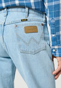 Un jean en denim bleu clair présente une patch logo Wrangler et une couture distinctive sur les poches arrière. Une chemise à carreaux bleue est visible au-dessus.