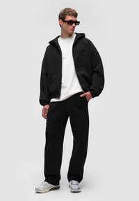 Felpa nera oversize e pantaloni abbinati in tessuto morbido, abbinati a una maglietta bianca e scarpe da ginnastica grigie. Gli accessori includono occhiali da sole neri.