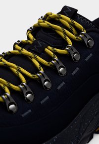 Must matkajalats, millel on kollase ja musta mustriga paelad, metallist silmad, tekstureeritud pealiskülg ja täpiline kummist tallas.