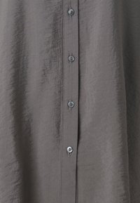 Chemise grise à boutons avec une texture froissée, dotée de six boutons gris sur le devant et d'une finition en tissu lisse.