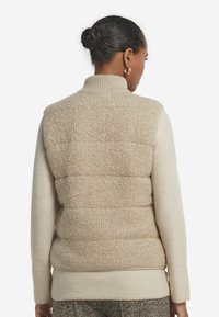 Beige vadderad väst gjord av mjukt, strukturerat tyg. Har en hög krage och lagerdesign, bärs över en krämfärgad tröja.