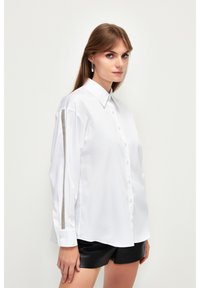 Camicia bianca con bottoni, a maniche lunghe e una texture liscia. Presenta un dettaglio a contrasto sulle maniche e un design classico del colletto.