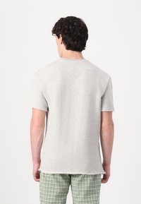 Tricou gri din bumbac, cu mâneci scurte, cu margini brute și decolteu rotund, purtat cu pantaloni în carouri verzi. Văzut din spate.