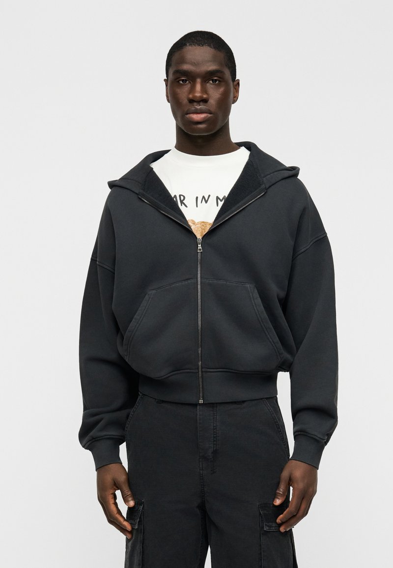 Μαύρο φούτερ με φερμουάρ, κατασκευ ασμένο από μαλακό ύφασμα, με μεγάλο κουκούλι, τσέπη καγκουρό και ριμπ μανσέτες· φορεμένο πάνω από ένα λευκό μπλουζάκι με γραφικό σχέδιο.