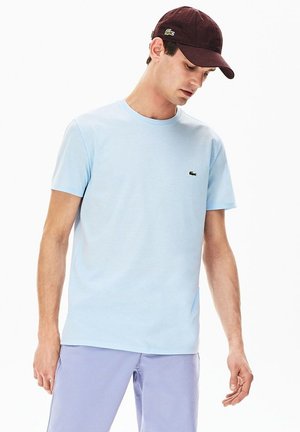 T-shirt en coton bleu clair avec un col rond et des manches courtes, arborant un petit logo vert Lacoste sur la poitrine. Assorti avec un pantalon lavande.